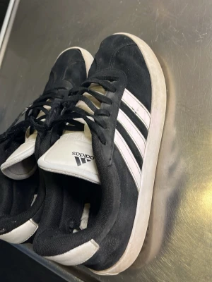 Svarta Adidas sneakers med vita ränder - Säljer ett par klassiska svarta Adidas sneakers med de ikoniska tre vita ränderna på sidan. Skorna har svart mockaliknande material, vita sulor och vit plös med Adidas-logga. Perfekta för en sportig och stilren look.