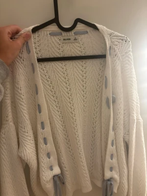 Vit stickad kofta från Hollister - Säljer en vit stickad kofta från Hollister med snyggt hålmönster och ljusblå banddetaljer längs kanterna. Koftan har långa ärmar och en avslappnad passform, perfekt att slänga över en topp. Materialet är mjukt och luftigt.