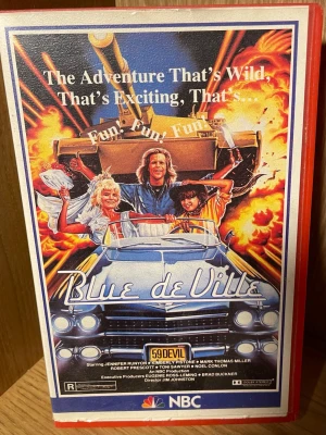 Blue de Ville - Actionkomedi VHS -  'Blue de Ville', en actionkomedi om två vänner på roadtrip i en klassisk Cadillac från 1959. Omslaget har färgstark illustration med explosioner, bil och karaktärer. Filmen är textad  är på svenska och utgiven av NBC/PRISMA FILM.