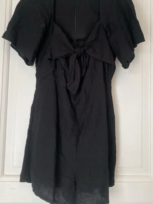 Svart jumpsuit med knyt och volangärm - Snygg svart jumpsuit med korta ben, volangärmar och en dekorativ knytning framtill. Dragkedja i ryggen för enkel påklädning. Tillverkad i ett lätt, vävt tyg som ger en avslappnad men trendig look.