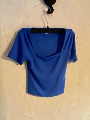 Blå topp från Amisu med vid urringning - Snygg blå topp från Amisu med korta ärmar och vid, något draperad urringning. Toppens material känns mjukt och stretchigt, perfekt till jeans eller kjol. Enkel men trendig design som passar till många olika stilar.