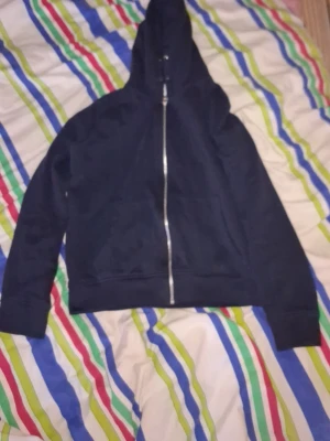 Svart hoodie med dragkedja - En enkel svart hoodie med dragkedja framtill och huva. Modellen har långa ärmar, breda muddar och en klassisk passform. Perfekt för en avslappnad stil och funkar till det mesta. Materialet känns mjukt och bekvämt, troligen bomullsblandning.