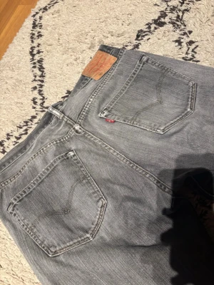 Grå Levi's jeans med raka ben - Säljer ett par grå jeans från Levi's med klassisk femficksmodell och raka ben. Jeansen har en snygg tvättad look, tydliga sömmar och den ikoniska läderlappen bak i midjan. Perfekta för en avslappnad stil och tillverkade i bomull.
