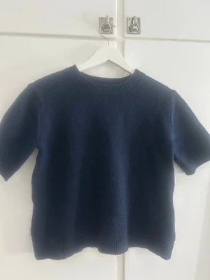 Mörkblå stickad tröja - En enkel och stilren mörkblå stickad tröja med rund halsringning och halvlånga ärmar. Tröjan har en rak passform och ribbstickade detaljer vid halsen. Perfekt att matcha med jeans eller kjol för en avslappnad look. Storlek 164 men passar xxs/xs