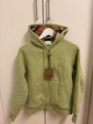 Ljusgrön hoodie från Burberry - Säljer en stilren och exklusiv Burberry zip-hoodie i färgen Mist Green. Klassisk modell med Burberrys ikoniska check-mönster i luvan och broderad logga framtill. Tillverkad i mjuk bomull av hög kvalitet.  ✔ 100 % äkta Burberry ✔ Storlek: S ✔ Färg: Mist Green ✔ Mycket fint skick ✔ Nypris: 8 900 kr  Perfekt hoodie som funkar både till vardag och mer uppklädda outfits. Skickas eller möts upp enligt överenskommelse.