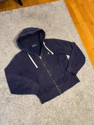 Äkta och mycket sökfunktion marinblå hoodie från Polo Ralph Lauren - Snygg marinblå hoodie med dragkedja från Polo Ralph Lauren. Tröjan har huva med vita snören, två fickor framtill och den klassiska gula loggan broderad på bröstet. Perfekt för en avslappnad och stilren look.