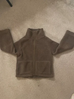 Brun teddyjacka med dragkedja - Mysig brun teddyjacka med hög krage och hel dragkedja framtill. Jackan har två stora fickor framtill och är tillverkad i fluffigt teddy-material som håller dig varm. Perfekt för kyliga dagar och enkel att matcha med olika outfits.