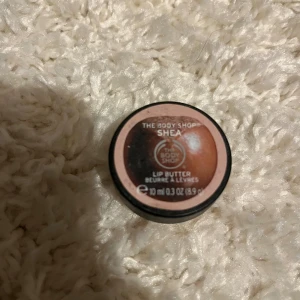Shea Lip Butter från The Body Shop - Shea Lip Butter från The Body Shop i en rund beige plastburk med skruvlock. Innehållet är ljusgult och har en krämig konsistens. Perfekt för att återfukta torra läppar med en mild doft av shea.