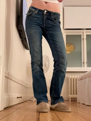 Blå bootcut jeans - Säljer ett par blå bootcut jeans med klassisk femficksdesign och låg midja. Jeansen har en snygg tvätt och lätt utsvängda ben. Perfekta för dig som gillar en retroinspirerad look och vill ha ett par jeans som sitter skönt och har en tidlös stil. Dom är köpta från Gina Young och är i storlek 170 men passar mig som har xs. Har fått ett litet hål på fickan som jag sytt igen