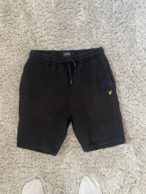 Svarta shorts från Lyle & Scott - Svarta mjukisshorts från Lyle & Scott med resårmidja och dragsko. Klassisk gul logga på vänster ben och två sidofickor. Perfekta för chill dagar eller träning. Tillverkade i mjukt sweatshirtmaterial för extra komfort.