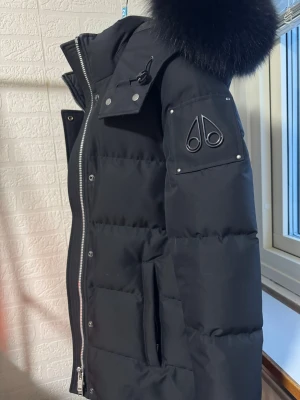 Svart dunjacka Moose Knuckles - Snygg svart dunjacka från Moose Knuckles med stor pälskantad huva och silverfärgade dragkedjor. Jackan har logotyp på ärmen, flera fickor och quiltad design. Perfekt för kalla vinterdagar och riktigt varm tack vare dunfyllningen. Använd fåtal gånger, nypris 12.000kr även köpt för 12.000kr… Tyvärr så har den blivit liten för mig och har en annan som jag använder mer.