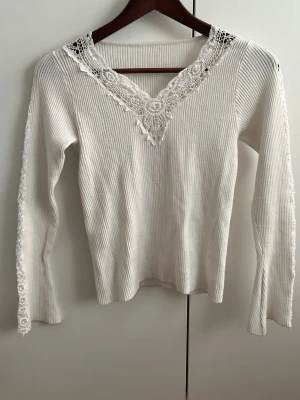 Vit ribbad topp med spetsdetaljer - Supersöt vit ribbad topp med lång ärm och v-ringning dekorerad med spets. Spetsdetaljer finns även längs axlar och ärmar för en extra feminin touch. Perfekt att styla med jeans eller kjol för en trendig look. Fits xs. Very good condition, no sign of use.
