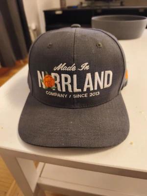Grå keps från SQRTN Company - Snygg grå keps från SQRTN Company med broderad text 'Made in Norrland' och en hjortron-detalj framtill. Kepsen har rak skärm och justerbar snapback baktill. Tillverkad i bomull och polyester, perfekt för dig som vill representera Norrland med stil.