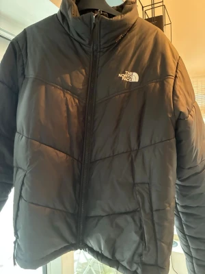 Svart pufferjacka från The North Face - Säljer en svart pufferjacka från The North Face med hög krage och dragkedja framtill. Jackan har diskret logga på bröstet och två sidofickor. Perfekt för kalla vinterdagar och riktigt skön att bära. Klassisk boxig passform och vadderad för extra värme.