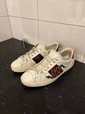 Gucci Ace  - Snygga vita sneakers från Gucci med klassiska gröna och röda ränder på sidan samt en broderad orm i rött och svart. Gucci ace orm (snake) Skorna har snörning och är tillverkade i skinn. Perfekta för dig som vill sticka ut med en ikonisk design.