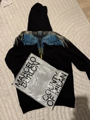 Marcelo Burlo M - Snygg svart hoodie från Marcelo Burlon County of Milan med det ikoniska blå vingtrycket över ryggen/axlarna. Högkvalitativ och stilren design som verkligen sticker ut. Bekväm passform och mjukt material, perfekt för både vardag och streetwear-outfits.Helt oanvänd då storleken ej passa