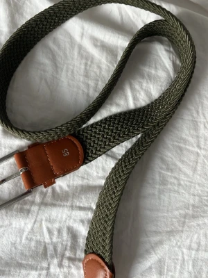 Olivgrönt flätat bälte med läderdetaljer - Snyggt olivgrönt flätat bälte med bruna läderdetaljer och silverfärgat spänne. Bältet har en flexibel design och passar perfekt till jeans eller chinos. Längd markerad som 85. Enkel och stilren accessoar som lyfter din outfit.