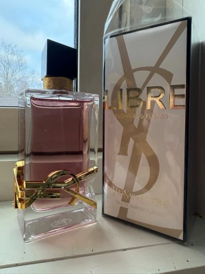 Yves Saint Laurent Libre Flowers & Flames - YSL Libre Flowers & Flames är en lyxig parfym i en stilren glasflaska med rosa doftvätska och guldiga detaljer. Flaskan har en svart, kantig kork och pryds av YSL:s ikoniska logga i metall. Kommer i originalförpackning med guldiga och svarta detaljer.