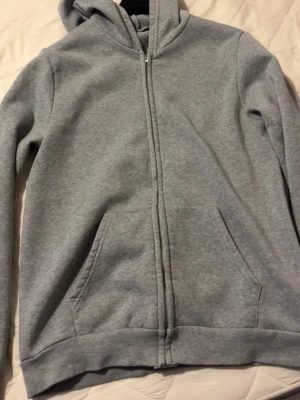 Grå zip-hoodie med fickor - En stilren grå hoodie med dragkedja framtill och två stora fickor. Hoodien har huva och ribbade muddar vid ärmslut och nederkant. Perfekt för en avslappnad look och enkel att matcha med andra plagg.