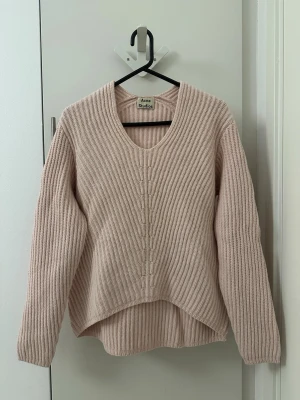 Ljusrosa stickad tröja Acne Studios - Säljer en ljusrosa stickad tröja från Acne Studios med v-ringning och ribbad struktur. Tröjan har lång ärm och något längre bakstycke för en avslappnad look. Perfekt för lager på lager och enkel att styla till olika outfits.