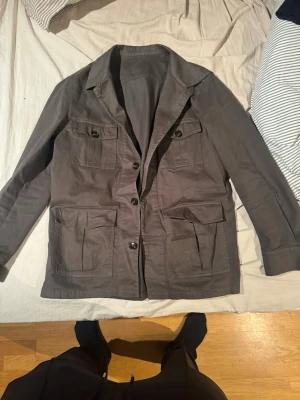 Grön safari jacka från Oscar Jacobson - Snygg grön Safari jacka från Oscar Jacobson med fyra stora fickor framtill och knäppning med svarta knappar. Jackan har klassisk krage och är tillverkad i ett slitstarkt bomullstyg. Perfekt för dig som gillar en stilren och funktionell look.