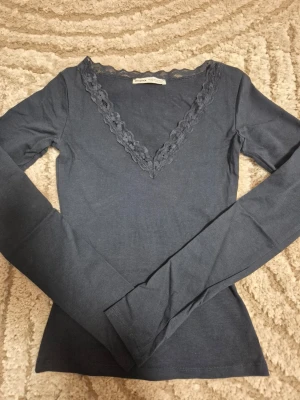 Mörkblå topp med spetsdetalj från bershka  - Supersnygg mörkblå långärmad topp från Bershka med v-ringning och spetskant runt halsen. Figurnära passform och mjukt material som känns skönt mot huden. Storlek xxs. Perfekt att styla med jeans eller kjol för en trendig look.