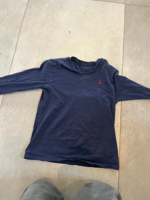 Mörkblå långärmad tröja Polo Ralph Lauren - Mörkblå långärmad tröja från Polo Ralph Lauren med klassisk rund hals och den ikoniska röda logotypen broderad på bröstet. Tillverkad i mjuk bomull som känns skön mot huden. Enkel och stilren design som passar till det mesta. Vanligt viss kostar den helt ny 800kr och jag har knappt använt den ni får mer en halva priset på den