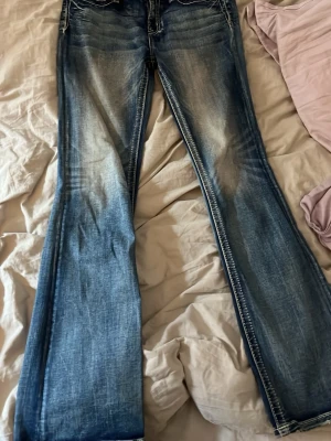 Blå bootcut jeans från Miss Me - Säljer ett par blå bootcut jeans från Miss Me med snygga slitningar och kontrastsömmar. Jeansen har låg midja och klassisk femficksmodell. En tråd är lite lös vid ena fickan 