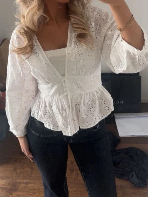 Vit broderad blus med knappar - Söt vit blus med broderade blommor och v-ringning. Blusen har dekorativa knappar framtill, volang nedtill och långa ärmar med lätt puff. Tillverkad i ett tunt, luftigt bomullstyg som ger en romantisk känsla. Perfekt att styla med jeans eller kjol.