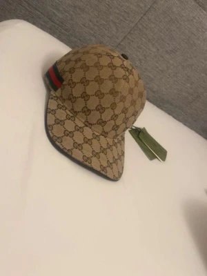 Gucci beige GG keps med logomönster - Säljer en beige keps från Gucci med det klassiska GG-monogrammet över hela kepsen. Kepsen har en böjd skärm, svart kant och en grön-röd rand på sidan. Tillverkad i canvasmaterial och har justerbar rem bak. Perfekt för dig som vill ha en ikonisk och trendig accessoar.