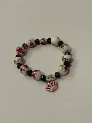 Blommigt pärlarmband med rosa berlock - Sött armband med vita pärlor dekorerade med rosa blommor och mörklila facetterade pärlor. Armbandet har en rosa berlock i form av en krona med glitterdetaljer. 