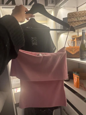 Rosa axelbandslös bandeautopp - Säljer en söt rosa bandeautopp med axelbandslös design och bred volang upptill. Toppen har en croppad passform och är perfekt för varma dagar. Materialet känns mjukt och stretchigt, vilket gör den bekväm att bära.