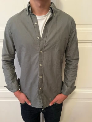 Grå skjorta från John Henric - Snygg grön/grå skjorta från John Henric med klassisk krage och knappar framtill. Skjortan har lång ärm och är tillverkad i mjuk bomull som känns skön mot huden. Perfekt att bära öppen över en t-shirt eller knäppt för en clean look.