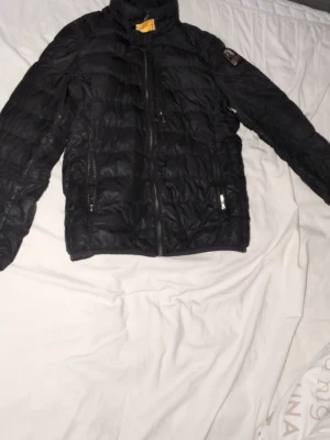 Svart dunjacka från Parajumpers - Snygg svart dunjacka från Parajumpers med klassisk patch på ärmen. Jackan har hög krage, dragkedja framtill och två fickor med dragkedja. Perfekt för kalla vinterdagar och riktigt skön att bära. Små hål på ärmen(ÄKTA)