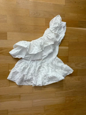Vit oneshoulder topp med volang - Säljer en superfin vit oneshoulder topp med stora volanger och broderade detaljer. Toppen är ärmlös och har en romantisk känsla med sin luftiga passform och broderade spetsmönster. Perfekt för dig som vill sticka ut lite extra!