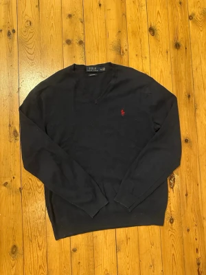 Mörkblå tröja från Polo Ralph Lauren - Klassisk mörkblå tröja från Polo Ralph Lauren med V-ringning och den ikoniska röda logotypen broderad på bröstet. Tröjan har långa ärmar och ribbade muddar vid ärmslut och nederkant. Perfekt för en stilren och tidlös look.