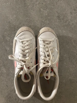 Vita Nike sneakers  - Säljer ett par klassiska vita Nike sneakers med rosa swoosh på sidorna. Skorna har lågt skaft, vita snören och detaljer i mocka vid tån och hälen. Perfekta för dig som gillar en clean och sportig look med en touch av färg.