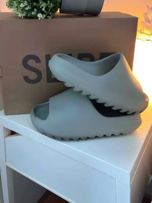 Gråa Yeezy Slide tofflor från Adidas - Säljer ett par Yeezy Slide tofflor från Adidas i en stilren grå färg. Tofflorna har en chunky, vågig sula och öppen tå för en modern look. Tillverkade i ett mjukt och flexibelt material som ger skön komfort. Perfekta för dig som gillar streetwear och minimalistisk stil.