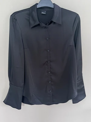 Svart satinskjorta med vida ärmar - Stilren svart blus i glansigt satinmaterial från Gina Tricot. Skjortkrage, knäppning framtill och långa vida ärmar med markerad manschett. Perfekt för dig som gillar en trendig och elegant look.