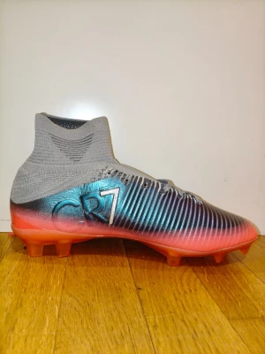 Nike CR7 Mercurial fotbollsskor - Snygga Nike Mercurial CR7 fotbollsskor med högt skaft i grått stickat material, metallicblå och svarta detaljer samt röd sula. Skorna har snörning och är designade för bästa grepp på planen. Perfekta för dig som vill sticka ut på matchen.