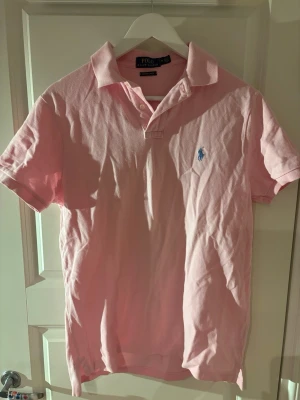 Ljusrosa pikétröja från Polo Ralph Lauren - Säljer en ljusrosa pikétröja från Polo Ralph Lauren med klassisk krage och korta ärmar. Tröjan har två knappar vid halsen och den ikoniska blå loggan broderad på bröstet. Perfekt för en avslappnad men ändå stilren look.