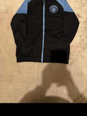 Manchester City svart fleece - Svart  med ljusblå detaljer och dragkedja. Jackan har Manchester Citys klubbmärke på bröstet och två fickor framtill.