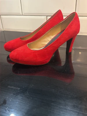 Röda mockapumps med klack - Snygga röda pumps i mocka med klassisk rund tå och hög klack. Skorna har en stilren design och innersula i beige. Perfekta för att lyfta vilken outfit som helst med sin djärva färg och eleganta form. Helt oanvända, bara testade och sen legat i en garderob