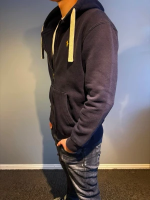 Ralph Lauren hoodie - Mörkblå hoodie från Ralph Lauren med dragkedja, vita snören och den klassiska gula loggan på bröstet. Den har storlek L. 