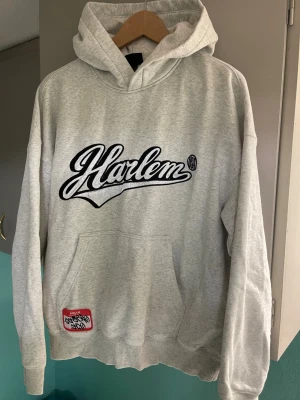 Grå Harlem hoodie med tryck - Snygg grå hoodie från Harlem med stor svart/vit logga på bröstet och tryck med siffran 86 samt romerska siffror på ryggen. Klassisk känguruficka framtill och huva. Tillverkad i mjukt bomullsmaterial, perfekt för en avslappnad streetwear-look.