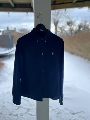 Ralph Lauren skjorta - marineblå - klassisk långärmad skjorta i bekvämt piqué-tyg (polo material). Marinblå med vit broderad logotyp. Mycket fint skick! • Storlek: XL (Boys 18–20 år). Motsvarar ca XS/S för herr eller S/M för dam. • Material: 100% bomull med skön stretch. Hör av dig vid frågor!🙂