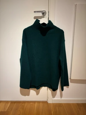 Mörkgrön stickad polotröja - Mysig mörkgrön stickad polotröja med hög krage och avslappnad passform. Tröjan har långa ärmar och är tillverkad i ett mjukt, tjockt material som passar perfekt för kyligare dagar. Enkel och stilren design utan synliga loggor eller mönster.