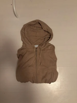 Beige hoodie med dragkedja - Säljer en beige hoodie med dragkedja framtill och justerbar huva med snören. Hoodien har långa ärmar och ribbade muddar vid ärmslut och nederkant. Perfekt för en avslappnad och stilren look.