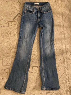 Blå bootcut jeans med vintagekänsla - Ett par klassiska blå bootcut jeans med lätt slitningar och tvättade detaljer. Jeansen har fem fickor, knappgylf och en avslappnad passform över benen. Perfekta för en avslappnad och trendig look.