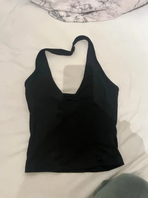 Svart halterneck linne - Säljer ett svart halterneck linne med djup urringning och smal rem runt nacken. Toppen är figurnära och har en enkel, stilren design utan synliga loggor eller detaljer. Perfekt att matcha med jeans eller kjol för en trendig look.